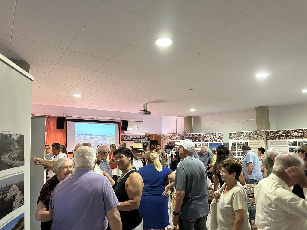 Fotografía de la sala de la exposición con varios asistentes, una pantalla al fondo con imágenes proyectadas y los roll-ups informativos de la muestra.