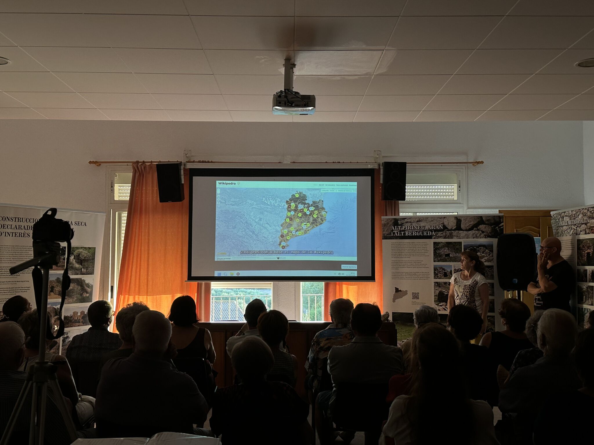Fotografía oscura con varios asistentes sentados observando un video proyectado en una pantalla central, donde se muestra fotograma del mapa de Cataluña con puntos marcados de construcciones de piedra seca, extraído de la web Wikipedra.