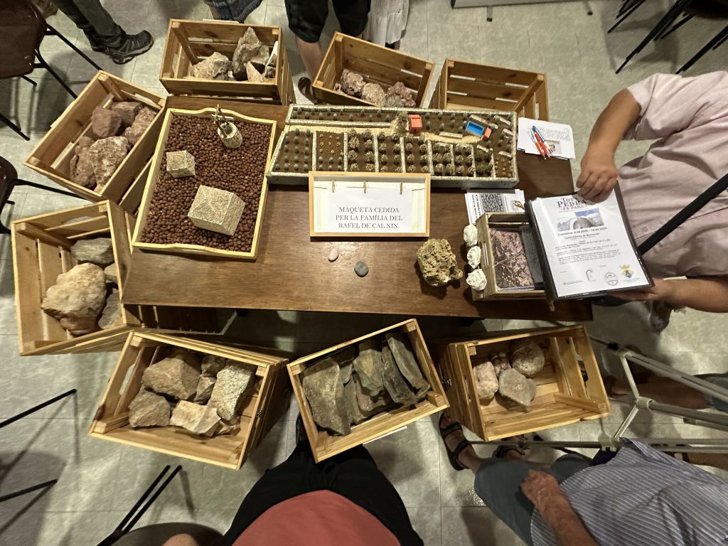 Fotografía de una mesa con dos maquetas hechas a mano: una representa un terreno y la otra un pozo con dos barracas de piedra seca. Sobre la mesa hay un dosier sobre las construcciones de piedra seca de La Pobla de Montornès y formularios para hacerse socio del Grup de la Pedra Seca de La Pobla de Montornès. Alrededor se ven cajas con distintos tipos de piedra para observar sus diferencias.